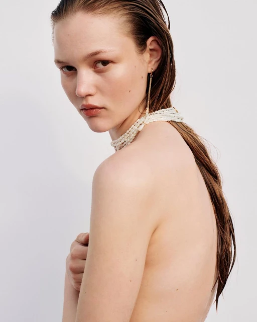 Portrait of a young woman with wet hair and a pearl choker, posing in front of a white background. She is looking at the camera.