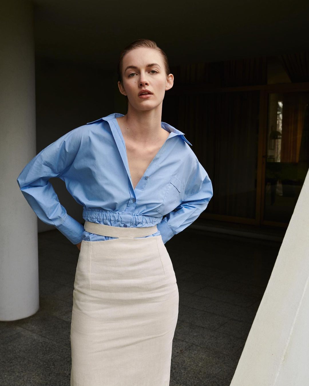 A fair-skinned woman poses in a blue collared shirt and beige skirt. The outfit is set against a muted indoor background with natural lighting.