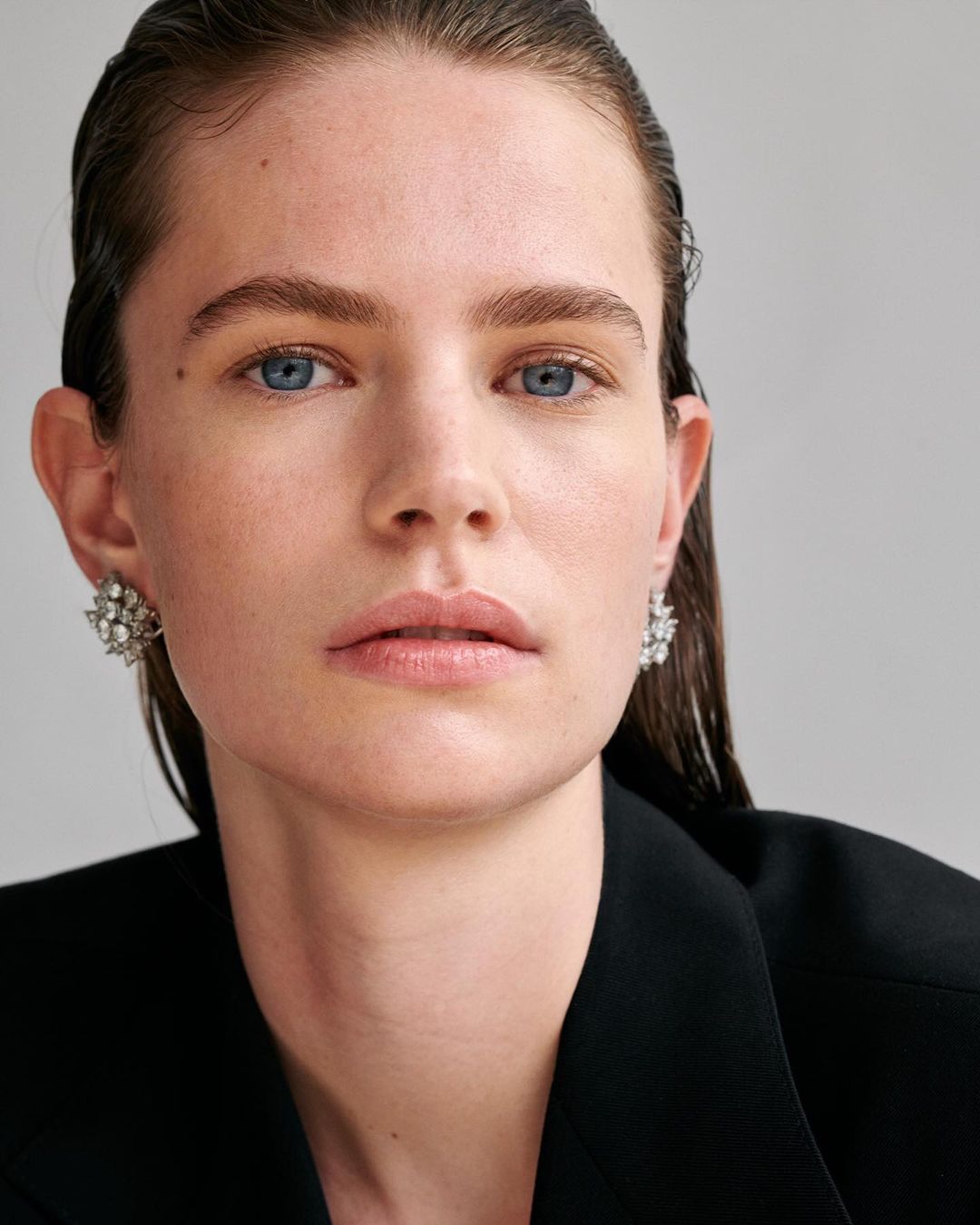 Close-up shot of a fair-skinned woman with slicked-back dark hair, wearing statement earrings, against a neutral gray background.