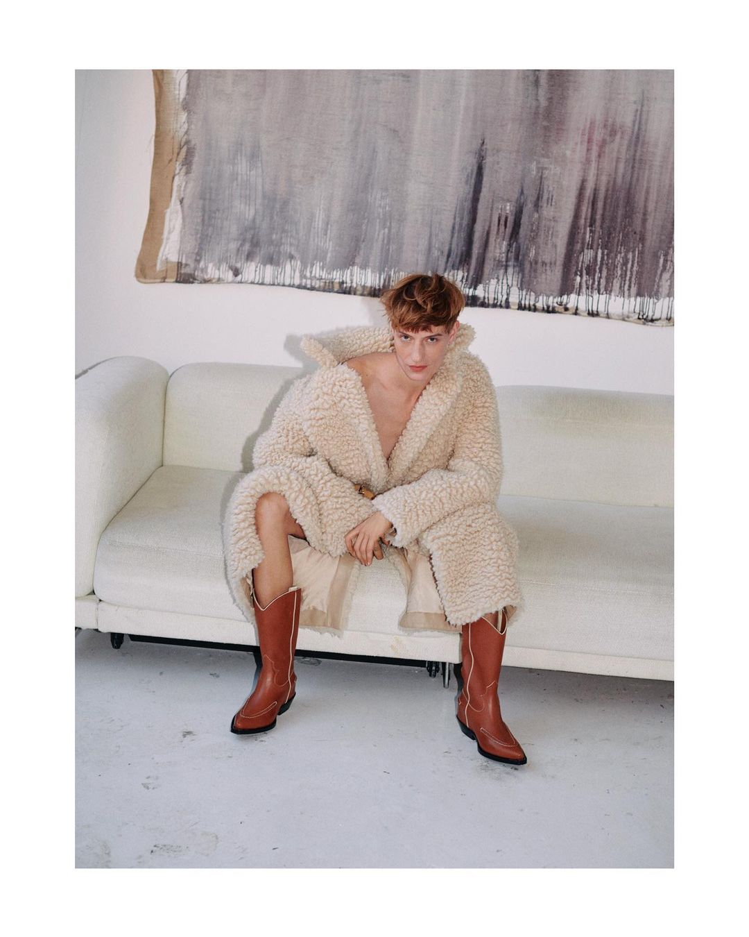 An adult male is seated on a white sofa wearing a cream-colored robe and brown cowboy boots in a well-lit indoor environment.