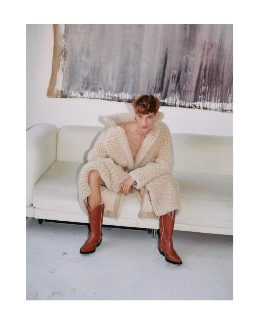An adult male is seated on a white sofa wearing a cream-colored robe and brown cowboy boots in a well-lit indoor environment.