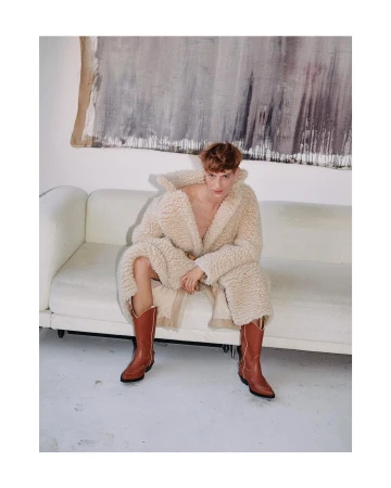 An adult male is seated on a white sofa wearing a cream-colored robe and brown cowboy boots in a well-lit indoor environment.