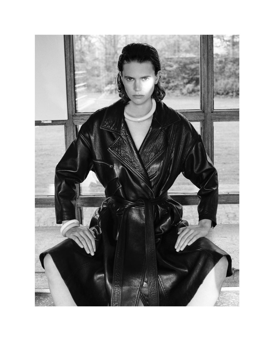 Monochrome portrait of a person in a black leather coat posing indoors by a window, showcasing fashion and style in a black and white aesthetic.