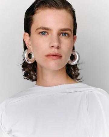 Close-up portrait of a woman with wet-look dark hair wearing large silver hoop earrings and a white top against a seamless white background.