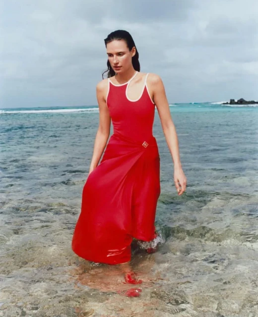Woman in a red dress walks through clear ocean water on a sunny day, with a bright sky and small island in the background.