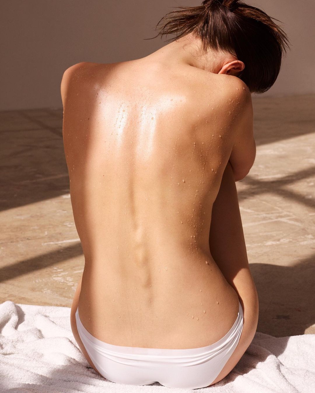 Back view of a wet, fair-skinned woman with tied-back hair sitting on a towel. The image features diffused light and visible water droplets.