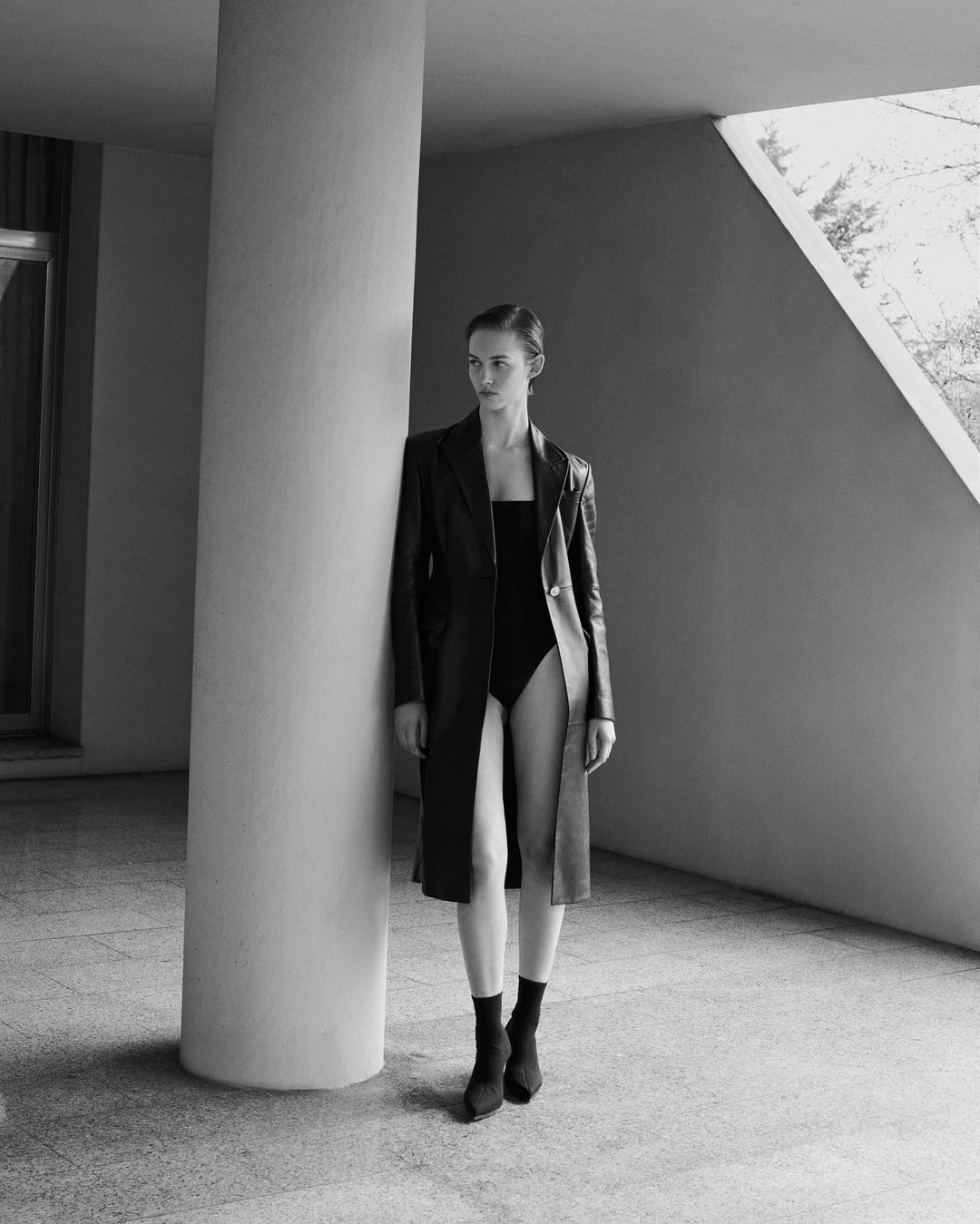 Monochrome portrait of a woman in a black bodysuit and coat, posing against a pillar in an architectural setting with natural light.
