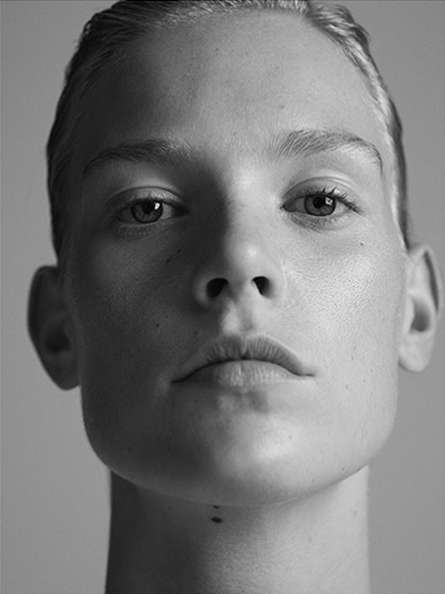 Black and white portrait of a young adult woman with slicked back hair, looking directly at the camera with a neutral expression.