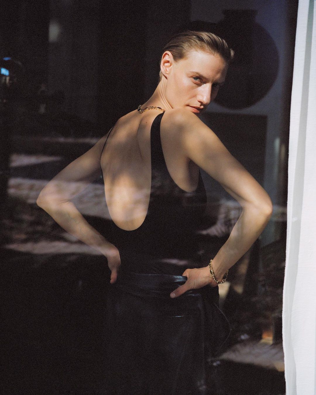 Woman in a black bodysuit and skirt poses in a softly lit indoor setting. Her hair is slicked back and she wears a gold bracelet and necklace.