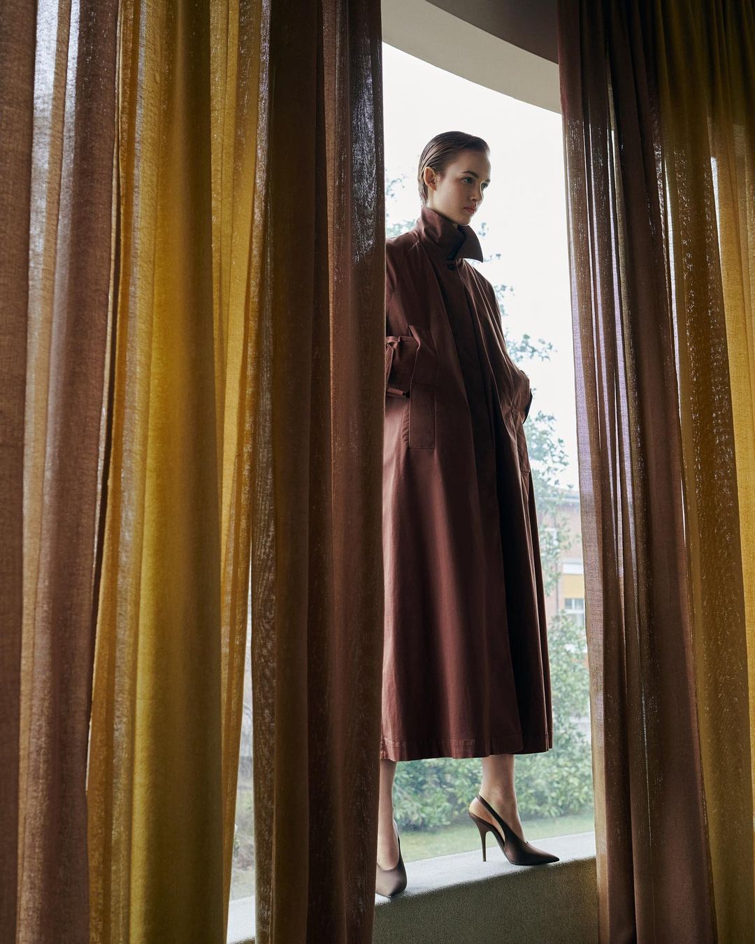 A woman in a brown coat and black high heels stands in a window framed by brown and yellow curtains. She is looking out the window.