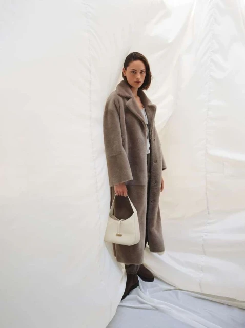 Fashionable woman poses in a full-length taupe coat, holding a cream handbag, against a draped white fabric backdrop in a studio setting.