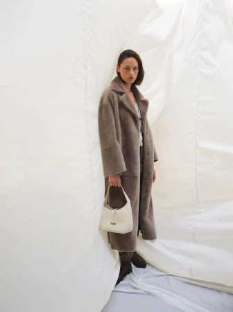 Model in grey fur coat and white handbag standing against draped white fabric backdrop photographed by Nadine Ottawa