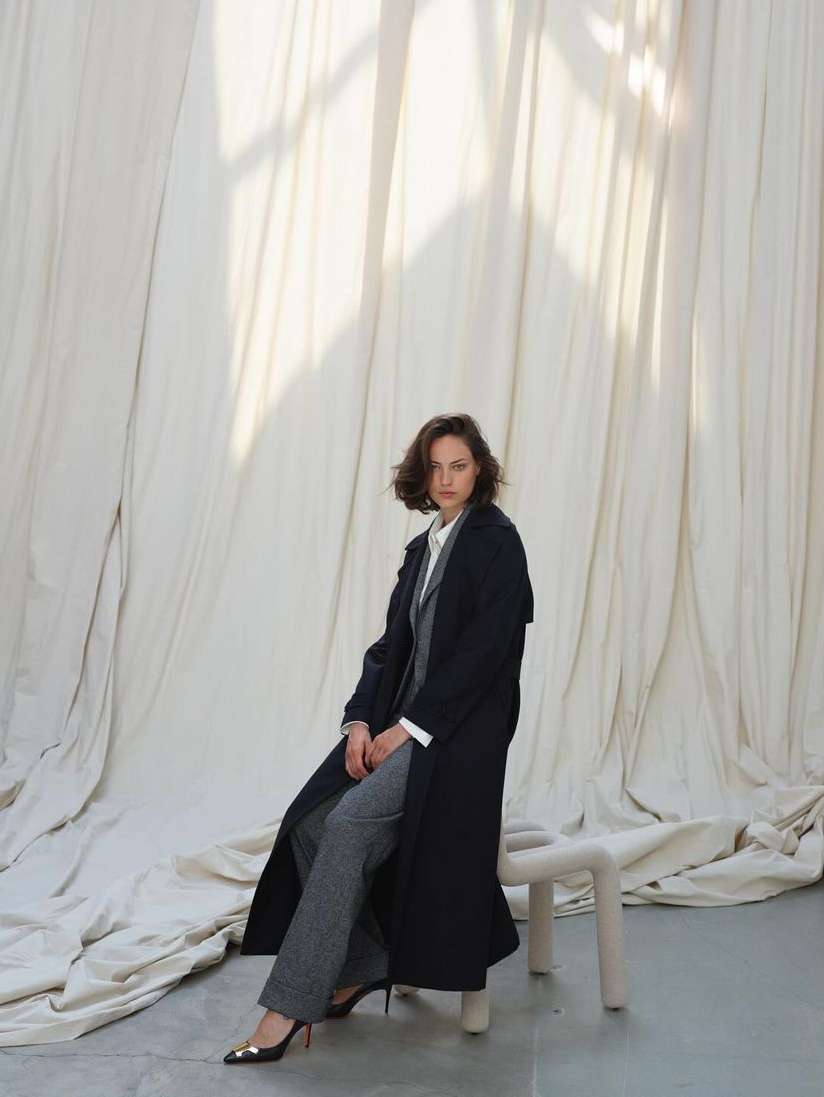 Woman in dark coat and grey trousers sitting on a stool against draped white fabric by photographer Nadine Ottawa