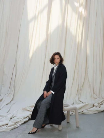 Woman in dark coat and grey trousers sitting on a stool against draped white fabric by photographer Nadine Ottawa