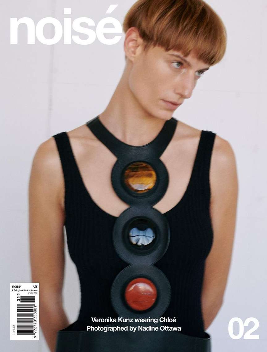 Portrait of a red-haired woman wearing a black top and large black circular necklace on a white background, magazine cover.