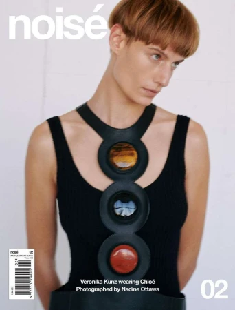 Model with short reddish hair wearing a black sleeveless top and a chunky statement necklace with circular gemstone insets against white.