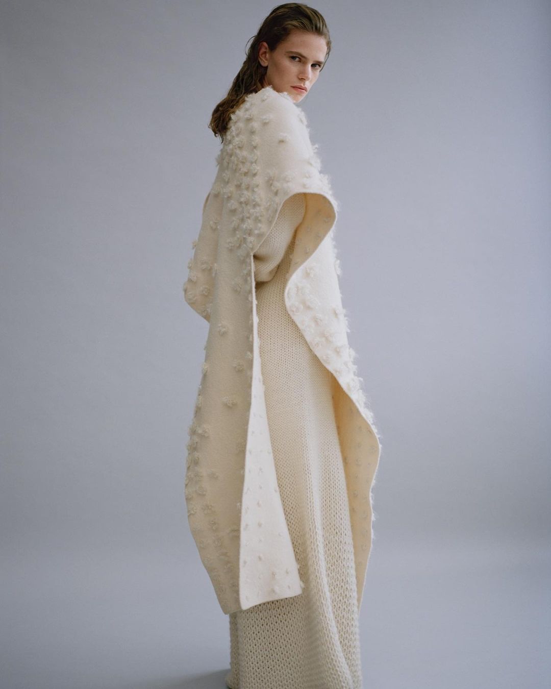 Woman in a full-length, cream-colored knitwear ensemble with oversized cape. She is looking over her shoulder in a studio setting.