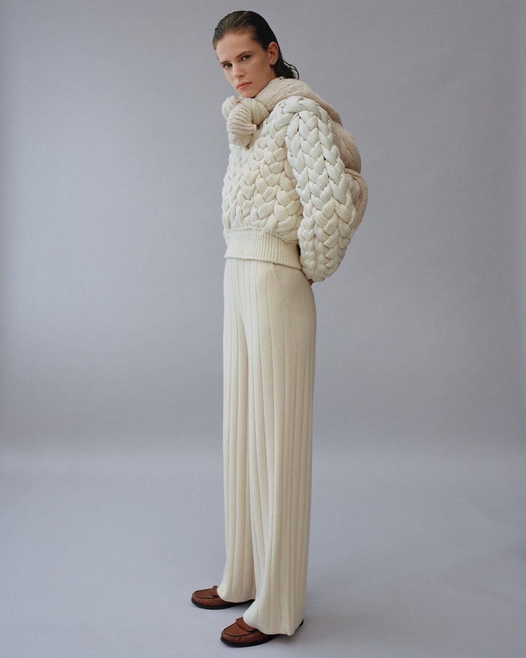 Full-length shot of a woman posing in a studio. She is wearing a cream-colored knit sweater, matching knit pants, and brown leather loafers.