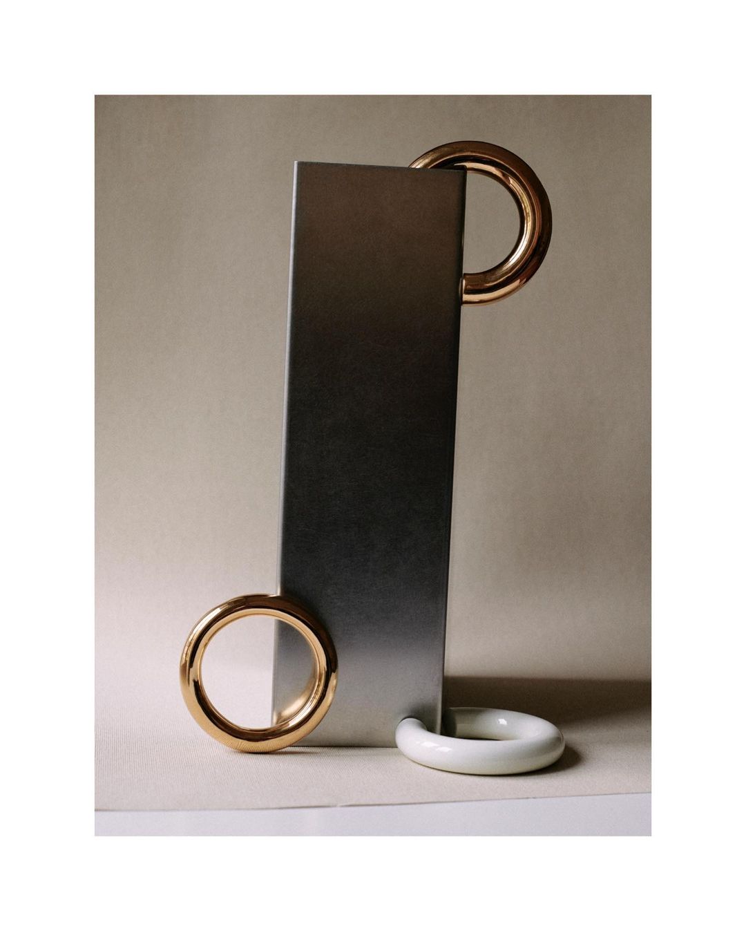 Abstract still life: A vertical metal bar is accessorized with golden and white rings on a beige background, creating geometric balance.