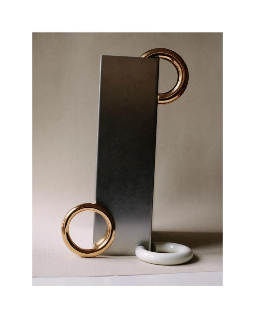 Abstract still life: A vertical metal bar is accessorized with golden and white rings on a beige background, creating geometric balance.