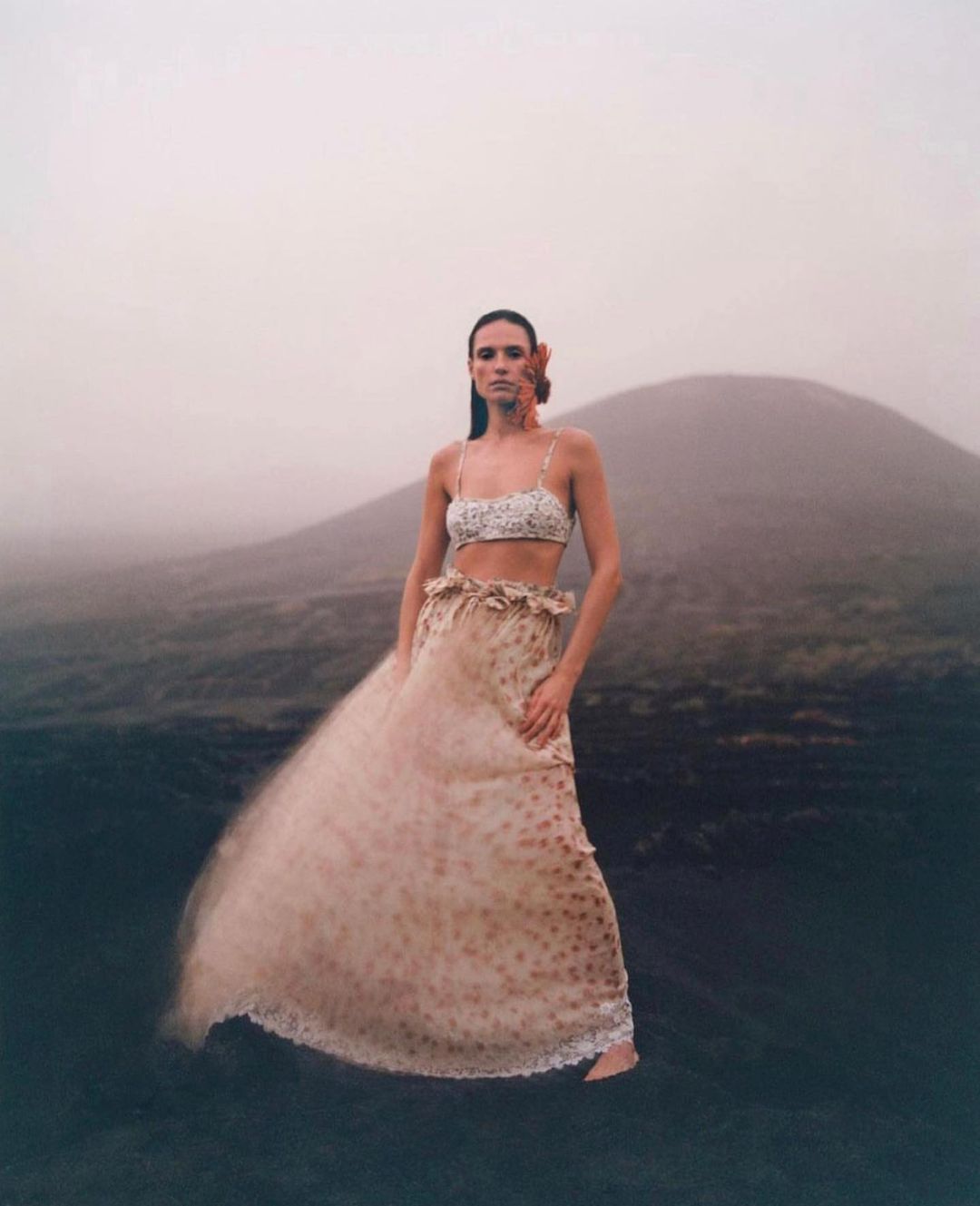 A woman in a flowing dress stands on black sand, with a flower behind her ear, set against a foggy background with a volcanic mountain.