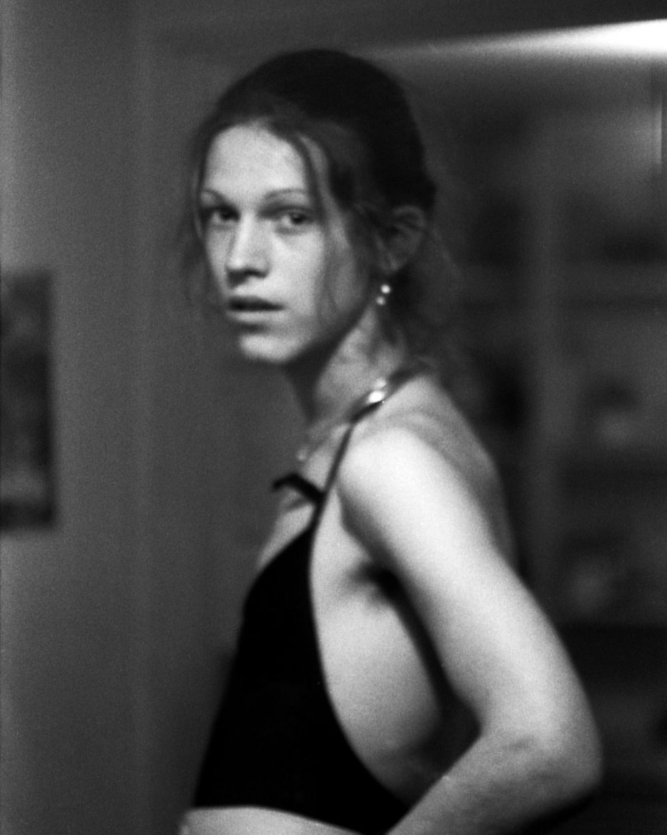 Monochromatic portrait of a young adult looking over their shoulder, featuring visible upper body and simple background.