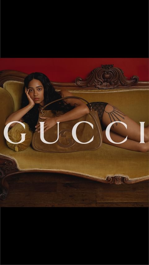 A woman lounges on an olive velvet couch with a brown suede Gucci bag, showcasing fashion and luxury in a warm-toned indoor setting.