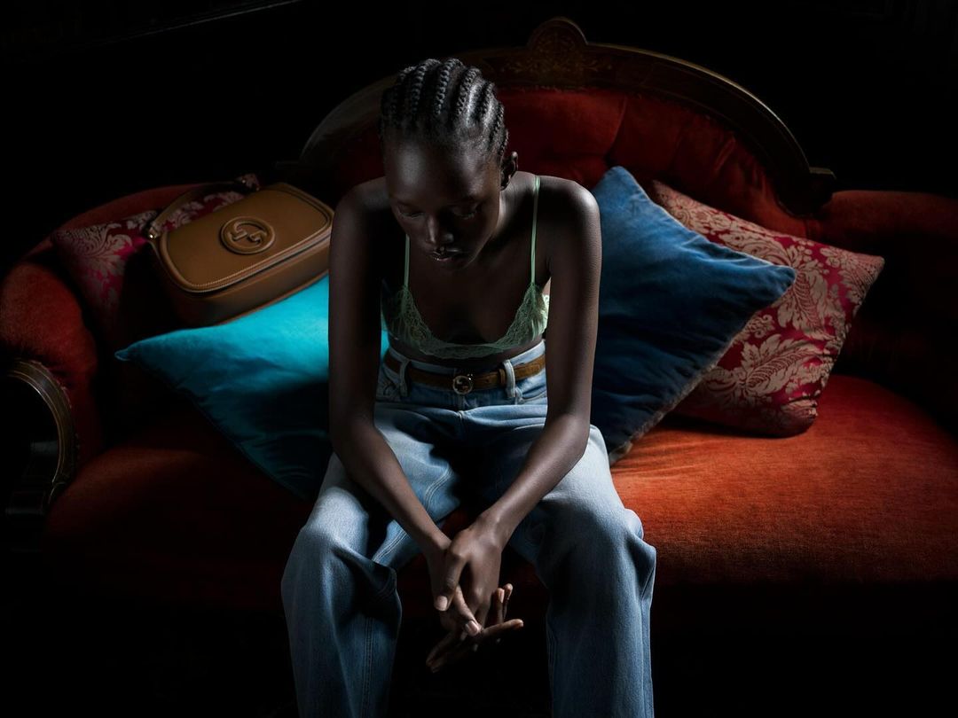 A seated, dark-skinned woman wearing a light-green top and jeans on a red velvet couch, accented by blue and patterned throw pillows.
