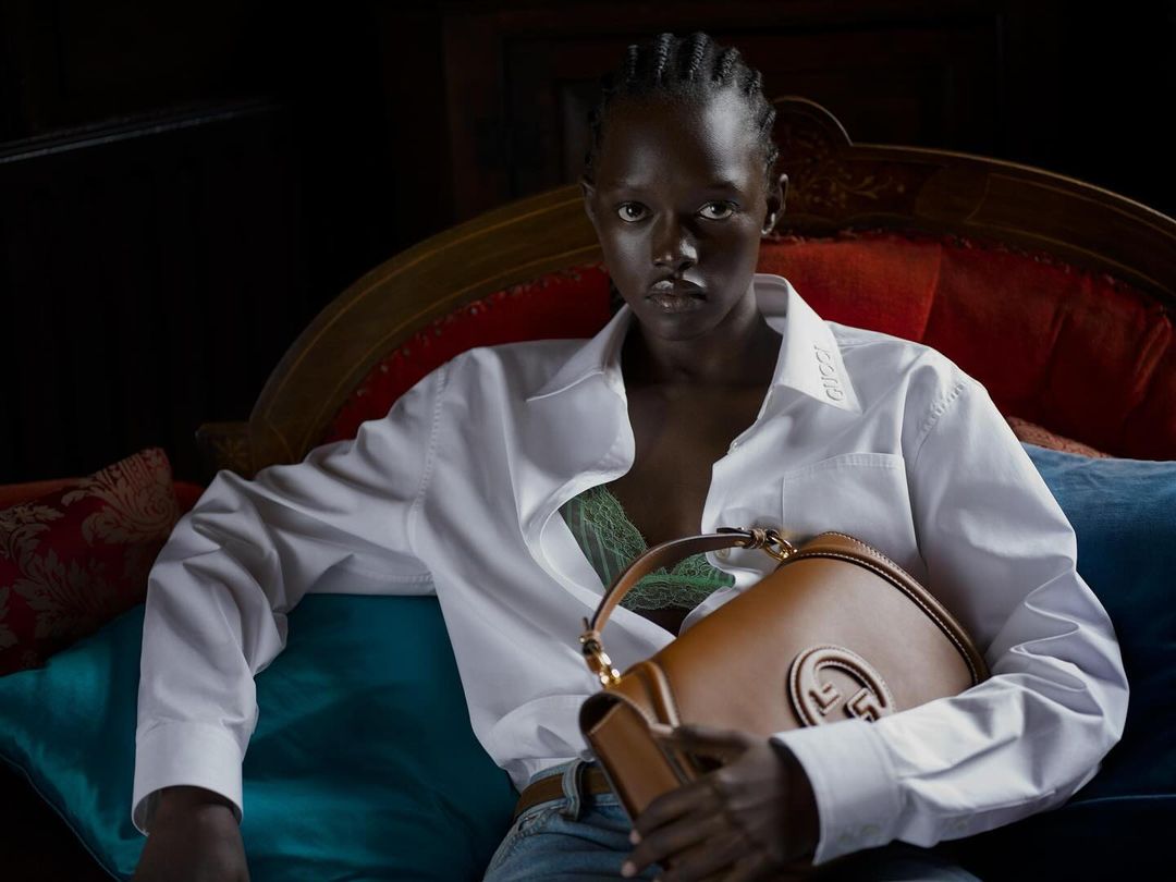 Elegant portrait of a Black woman in a white shirt, posing on a red and blue vintage sofa with a brown leather handbag.