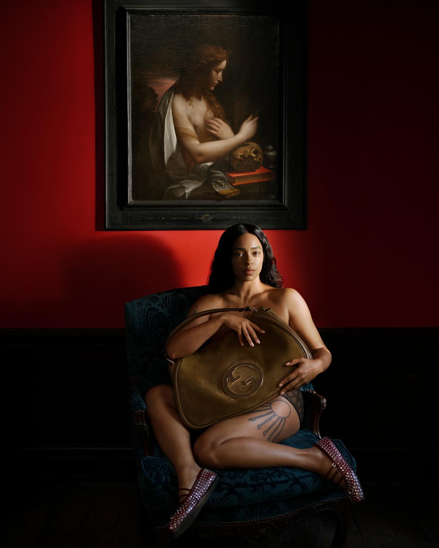 Nude woman sitting on chair holds gold Gucci bag in an art gallery with a classic painting and red walls in a medium studio shot.