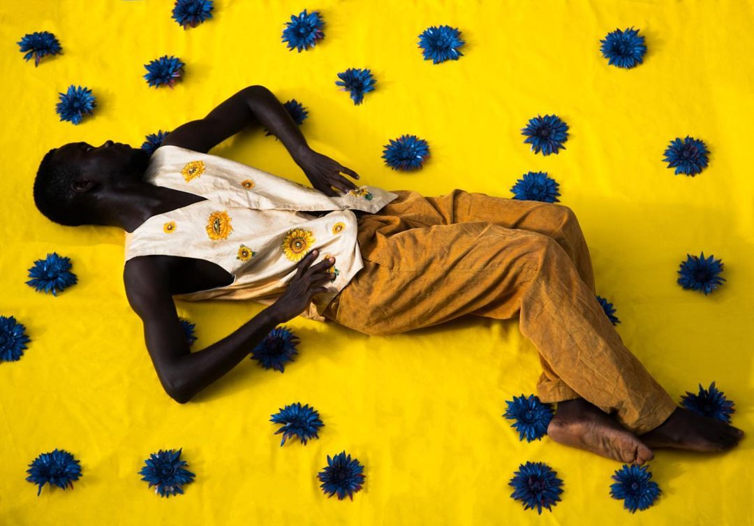 An African American man in a floral vest and gold pants is lying on a yellow surface covered with blue cornflowers in a studio shot.