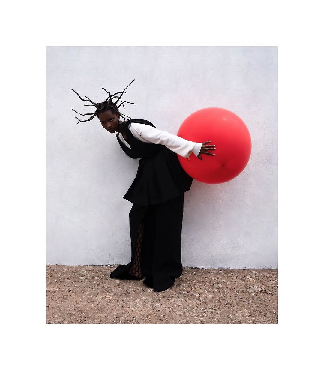 Fashionable Black woman in black and white outfit leans back against white wall while holding a large red balloon in a studio setting.