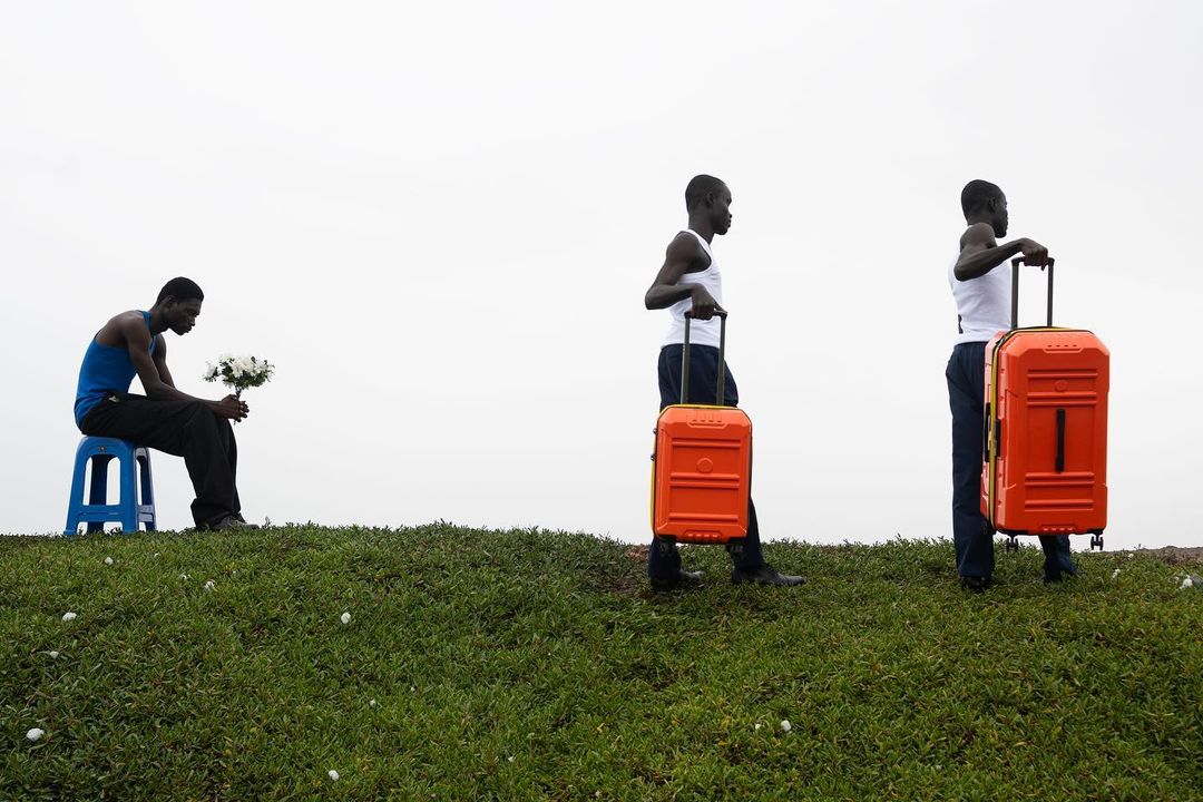 Three African men on grassy hill: one sits with flowers, two stand with orange suitcases, set against a bright, overcast sky.