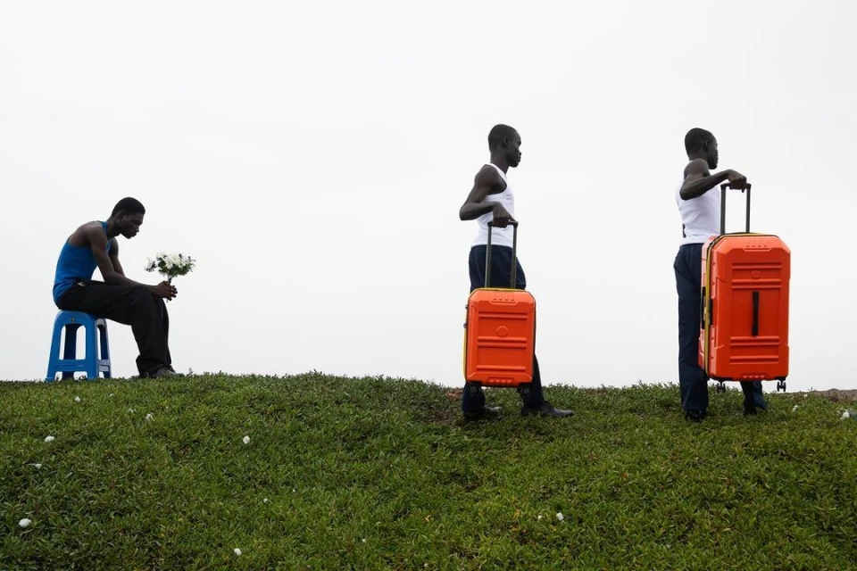 Three African men on grassy hill: one sits with flowers, two stand with orange suitcases, set against a bright, overcast sky.