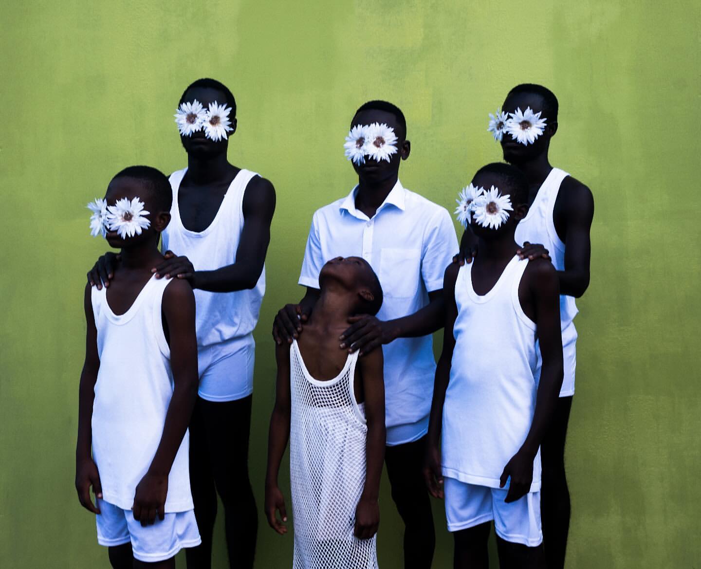 Conceptual fashion shot of a group of dark-skinned children and young adults in white clothing posing against a green-yellow wall, faces obscured with daisies.