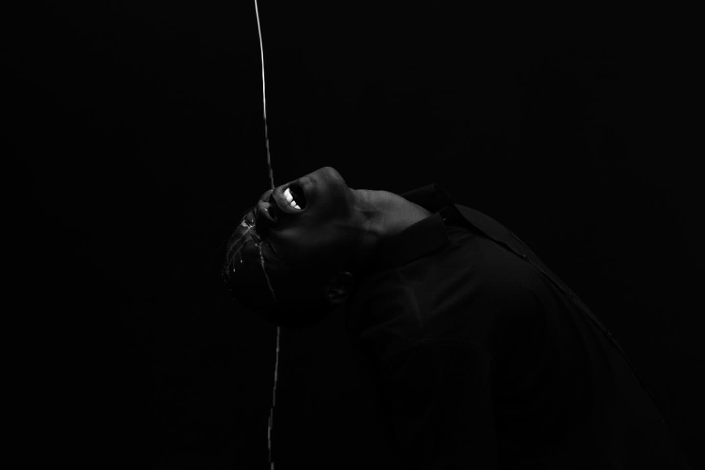 Monochrome image of adult with open mouth in dark environment with water streaming down face and torso. Liquid motion photography.