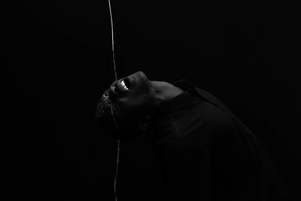 Monochrome image of adult with open mouth in dark environment with water streaming down face and torso. Liquid motion photography.