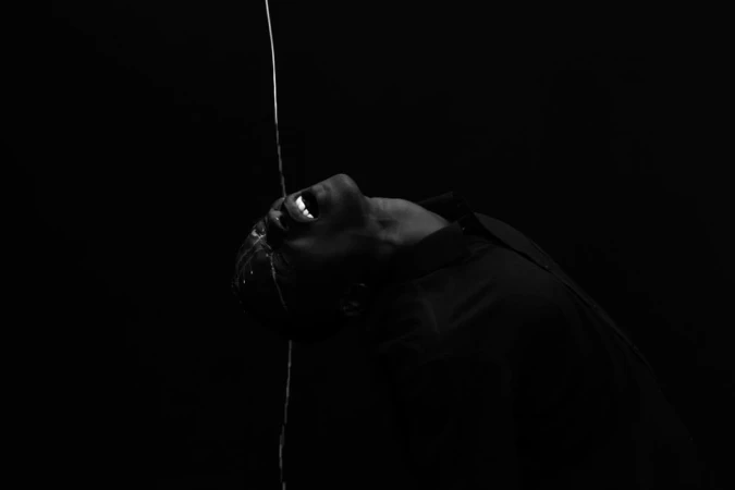Monochrome image of adult with open mouth in dark environment with water streaming down face and torso. Liquid motion photography.