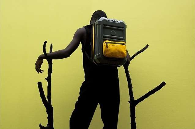 Man in black attire carries a unique gray backpack with yellow pocket, beside dark branches, set against a muted yellow backdrop.