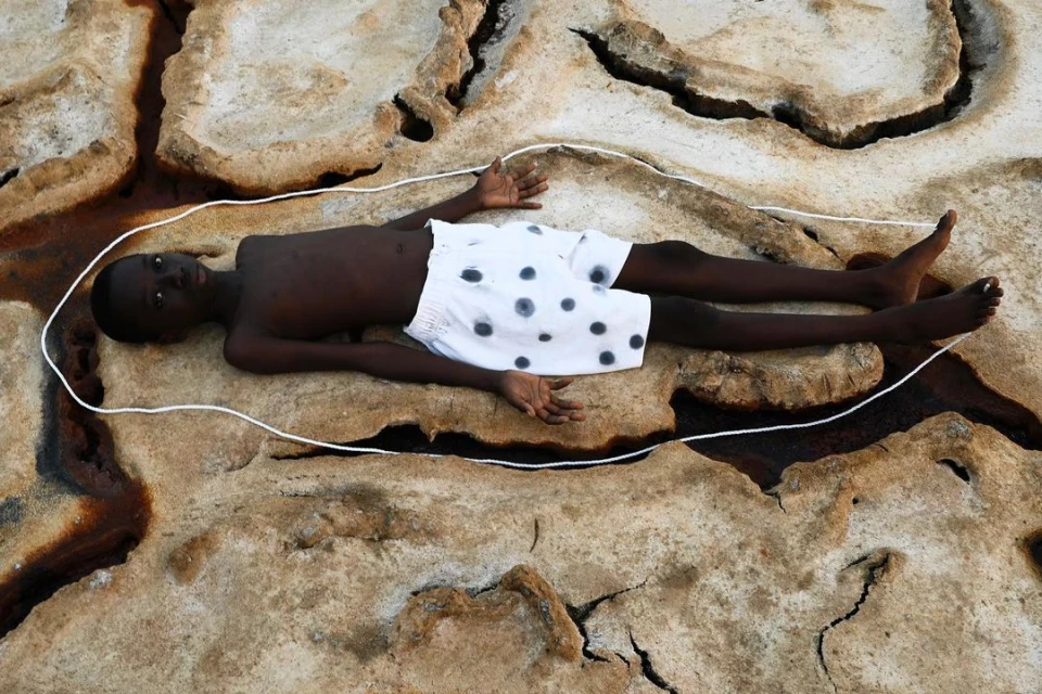 Child lies on cracked, light brown earth with a white rope outline, wearing patterned white shorts, with rust-colored liquid visible.