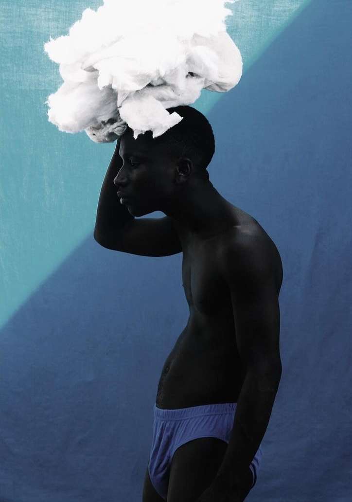 Studio shot of a Black male model with bare torso and cloud-like cotton on his head against a blue background. Artistic portrait.