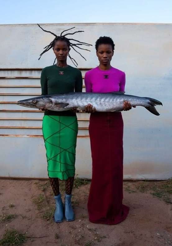 Two women in long skirts hold a large fish in front of a white wall, under a bright sky. Outdoor fashion portrait. 