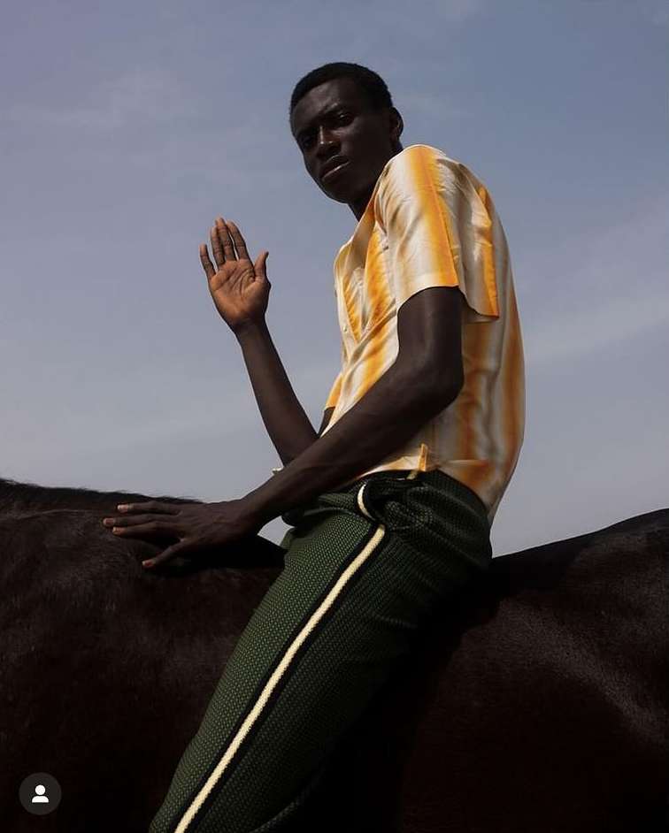 A man sits bareback on a black horse outdoors, gazing at the camera with a raised hand, wearing a yellow striped shirt and green pants.