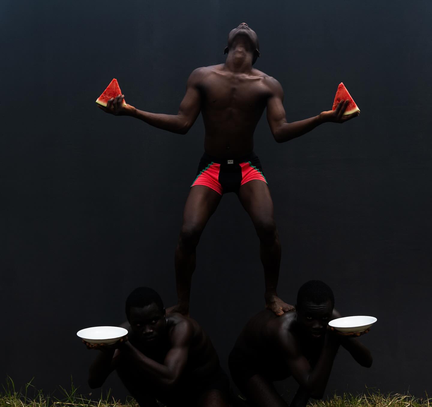 Three Black men in artistic pose: two supporting a third holding watermelon slices against a dark background with grass in the foreground.