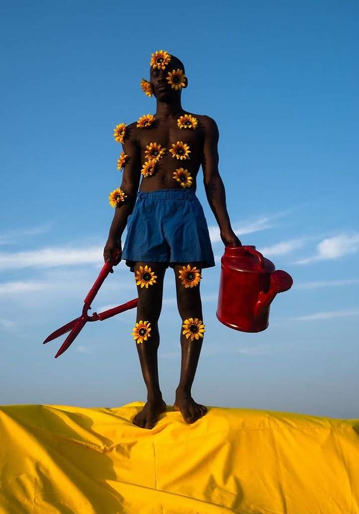 Adult male stands on yellow material against a blue sky, adorned with sunflower heads and holding a red watering can and shears.
