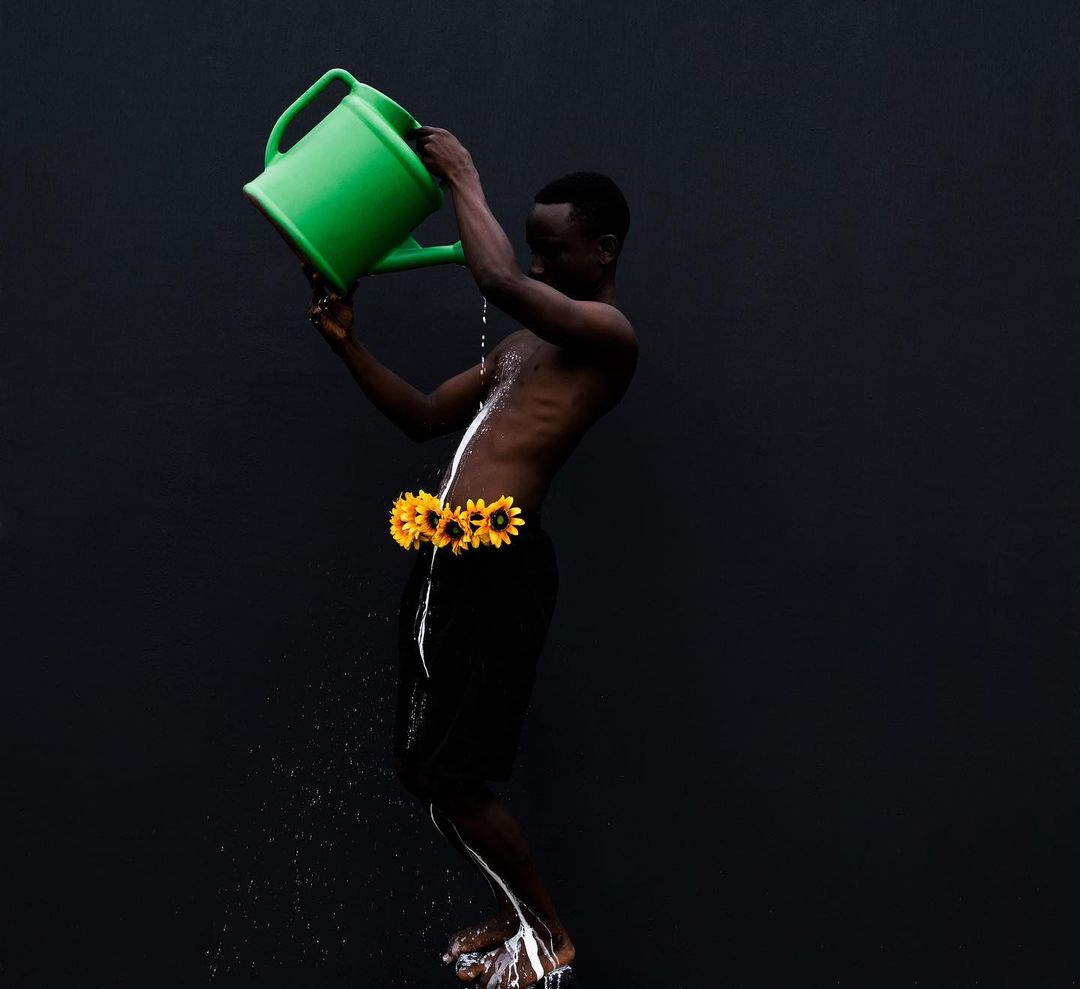 An African male watering his torso with a green watering can while wearing sunflowers in front of a black studio background.