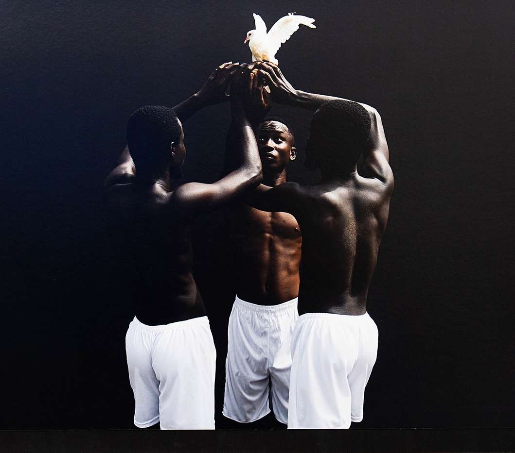 Three shirtless Black men in white shorts converge around a white dove against a dark background, creating a striking, artistic studio portrait.