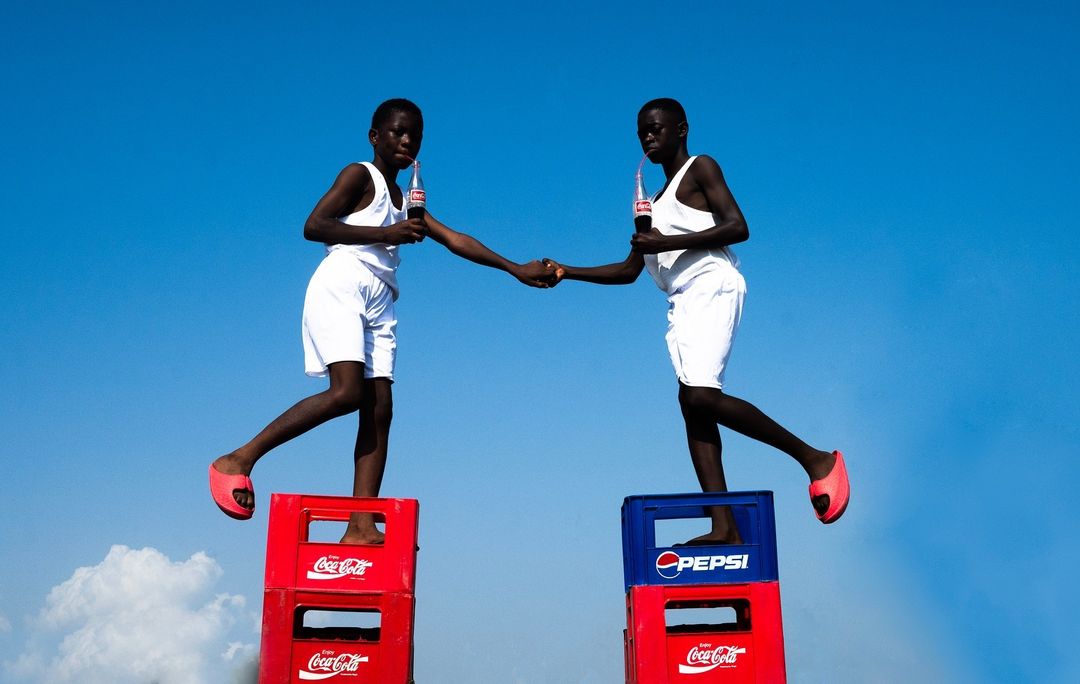 Two young individuals stand atop stacked beverage crates, bottles in hand, against a clear blue sky, creating a striking and balanced composition.