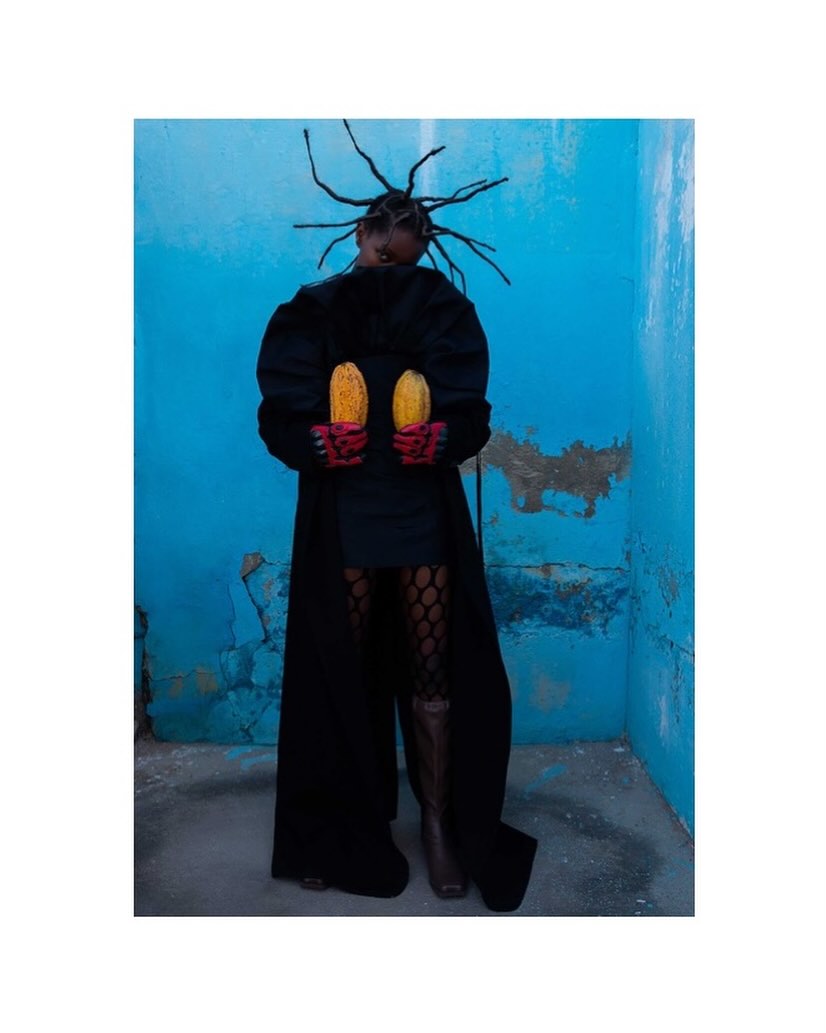 A striking studio portrait of an African woman in a black coat, holding corn cobs against a textured blue wall, showcasing unique fashion.