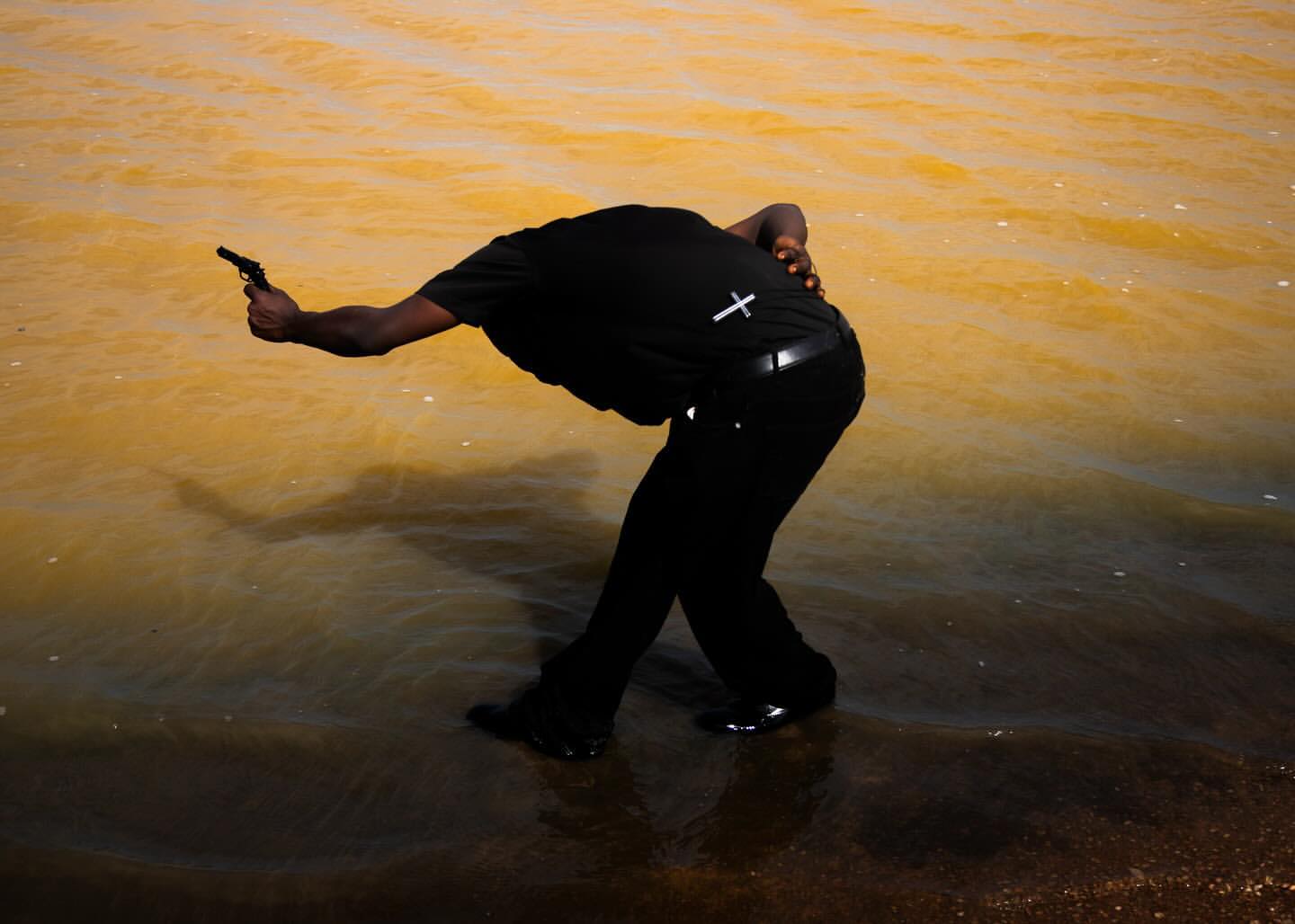 Adult male stands in shallow water with a small handgun, wearing all black with a small cross on his back. 